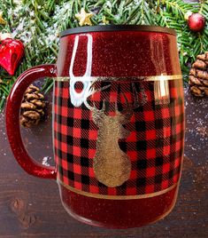 a red and black plaid coffee mug with a moose on it next to pine cones