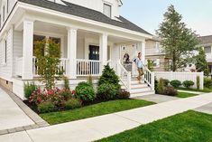 a woman is standing on the front porch of a white house with flowers and shrubs