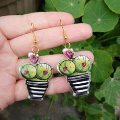 a pair of green cactus earrings with pink hearts on them sitting in front of a plant