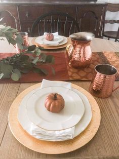 a wooden table topped with white plates and copper cups filled with pumpkins on top of them