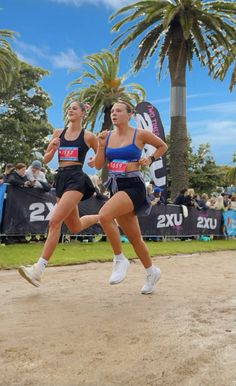 two women are running in the dirt near palm trees and people watching from behind them