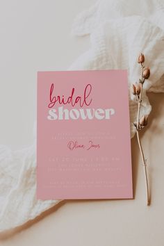 a pink bridal shower card sitting on top of a white blanket next to a flower