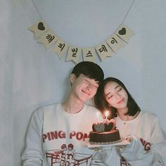 two people standing next to each other holding a cake with lit candles on it in front of a bunting banner