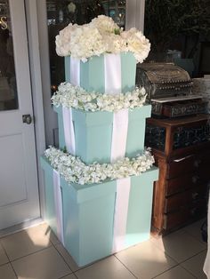 a three tiered cake decorated with white flowers and blue boxes sitting on the floor in front of a door