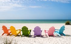 four colorful lawn chairs sitting on top of a sandy beach next to the ocean and blue sky