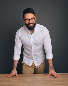 a man with glasses and a beard standing at a table in front of a gray background