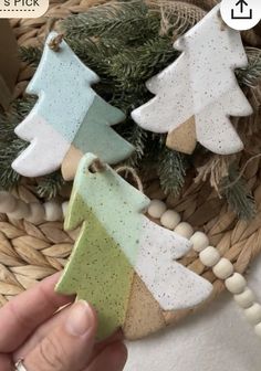 hand holding small ceramic christmas tree ornaments in front of a basket filled with pine branches