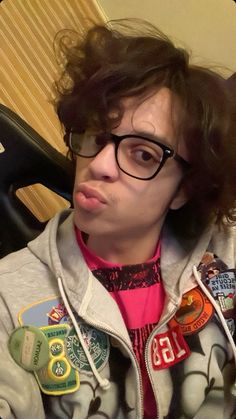 a person wearing glasses and a hoodie with patches on it's chest, making a funny face