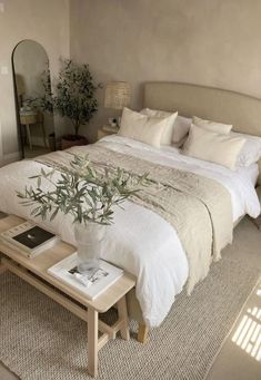 a bed with white linens and pillows in a bedroom next to a plant on a coffee table