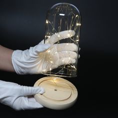 a person in white gloves holding a jar with string lights on it and a wooden lid