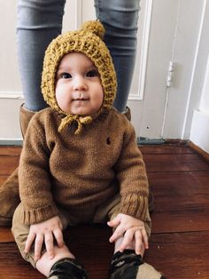 a baby sitting on the floor wearing a knitted hat and sweater with ear flaps