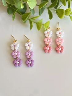 three pairs of pink, white and purple flower earrings on top of a table next to green leaves