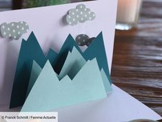 a handmade card with mountains and clouds on it, sitting on a table next to a candle