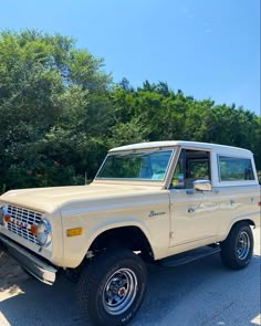 an old ford bronco is parked in a parking lot with trees and bushes behind it