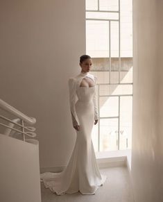 a woman standing in front of a window wearing a long sleeved wedding dress with an open back