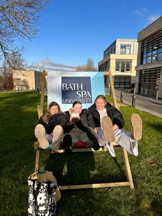 three women sitting on a wooden chair in the grass with a bath spa sign behind them