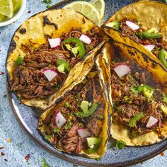 three tacos on a plate with limes and cilantro