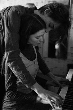 a man and woman are playing the piano in black and white, one is holding his head over the other's shoulder