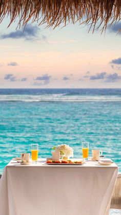 two glasses of orange juice sit on an outdoor table overlooking the ocean with straw umbrellas