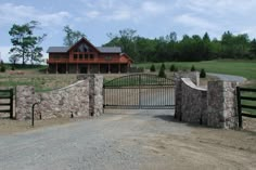 a stone fence and gate in front of a large house on a dirt road with trees around it