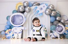 a baby sitting on the floor in front of balloons and space suits, smiling at the camera