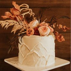 a white cake with flowers and greenery on top sitting on a wooden table in front of a wood wall