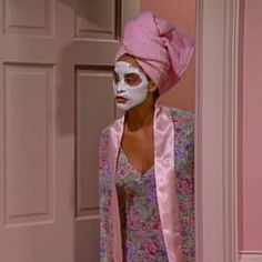 a woman with white face paint and a pink towel on her head standing in front of a door