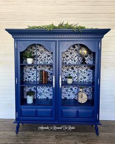 a blue china cabinet with plants and books on it's glass doors, in front of a white wall