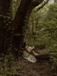 a woman sitting under a tree in the woods reading a book while wearing a white dress