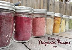 seven jars filled with different colored powders on a wooden table next to a fence