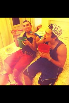 two young men sitting on a bed eating ice cream and drinking from plastic cups in front of them