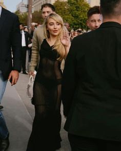 a woman in a black dress walking down the street with other people behind her and onlookers