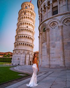 a beautiful woman in a white dress standing next to a tall tower with a clock on it's side