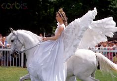 a woman dressed as an angel rides a white horse in a grassy area with people watching