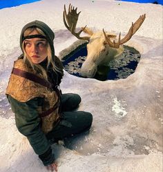 a woman kneeling in the snow next to a fake moose's head and water hole