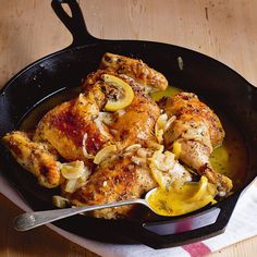 a skillet filled with chicken and lemons on top of a wooden table next to a fork