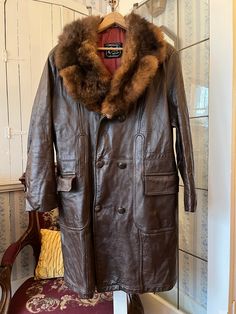 This gorgeous coat from Craft of Canada is made from genuine leather in dark brown, with a thick, soft collar of possum fur in natural medium to deep brown tones. It has the original leather covered buttons in the front and on the cuffs, two front pockets and one inside pocket, and it's fully lined with brown quilted fabric on top and red, beige and black 85 per cent wool lining on the bottom. The measurements, taken with the coat lying flat, are: shoulder to shoulder, 19 inches; armpit to armpit, 23 inches; sleeves, 25 inches; length, 42 1/2 inches; bottom edge, 31 inches. In very good condition. Brown Overcoat, Brown Leather Coat, Leather Trench, Brown Fur, Quilted Fabric, Leather Trench Coat, Brown Tones, Deep Brown, Fur Collar