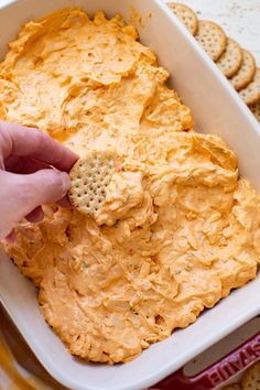 a hand dipping crackers into a white dish filled with hummus and cheeses