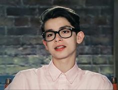 a young man wearing glasses sitting in front of a brick wall and looking at the camera