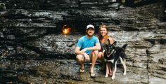 a man and woman sitting next to a dog in front of a fire place on the rocks