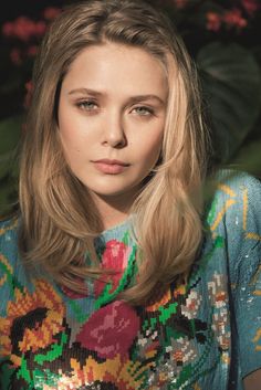 a woman with long blonde hair wearing a colorful sweater and holding a pink object in front of her face