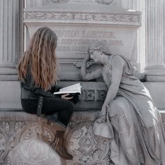 a woman sitting on the side of a building next to a statue holding a book