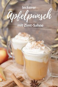 two mugs filled with coffee and topped with whipped cream next to cinnamon sticks, an apple