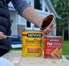 a person holding a paint can next to two cans of wood stain and a spoon