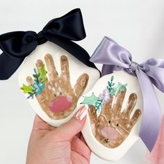 two handprinted ornaments are being held up by someone's hands with a bow