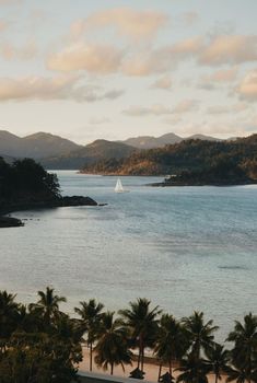 a sailboat on the water near some palm trees and mountains with clouds in the sky