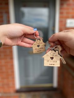 two hands holding wooden house shaped keychains in front of a brick building with the door open