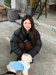 a woman kneeling down next to a small white dog wearing a blue and black jacket