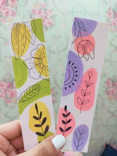 two handmade bookmarks with flowers and leaves painted on them are held in front of a floral wallpaper