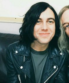 two people sitting on a couch smiling at the camera with one person wearing a leather jacket